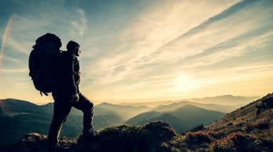 The man standing with a camping backpack on a rock with a picturesque sunset