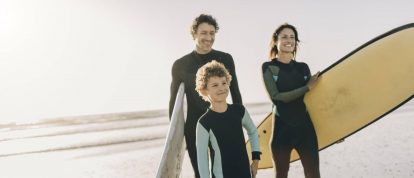 Lächelnde Familie, die im Frühling mit Surfbrettern am Strand entlang spaziert, Campingurlaub Angebote im April