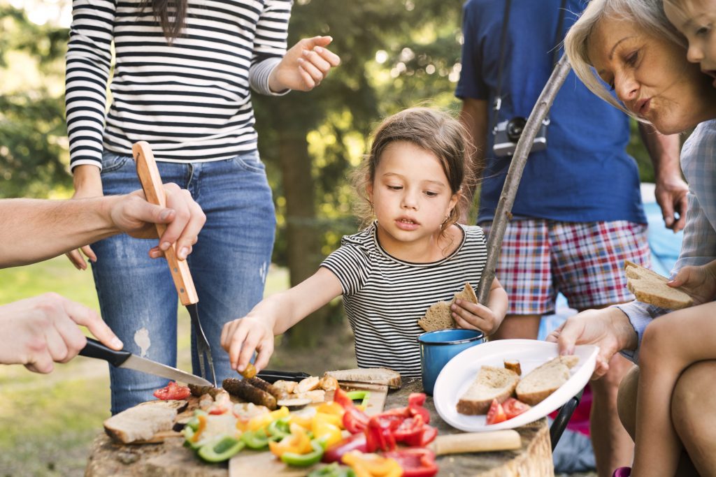 Kleines Mädchen greift über einen mit Essen bedeckten Tisch beim Camping mit der Familie