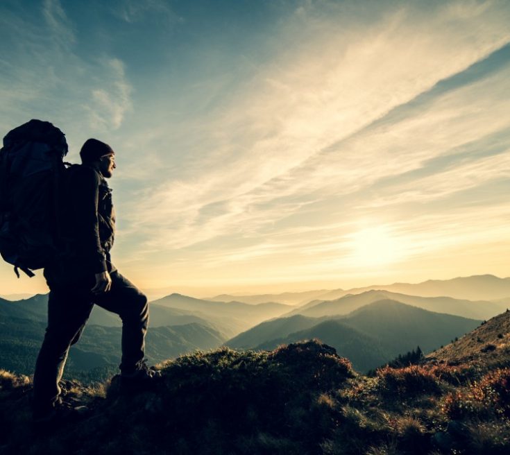 Mann mit Backpack betrachtet den Sonnenuntergang über einem Bergpanorama - Camping Checkliste
