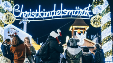 Weihnachtsmarktbesucher vor dem Weihnachtsmarkt in Straßburg, im Hintergrund weihnachtliche Beleuchtung und das Christkindlesmärik-Schild