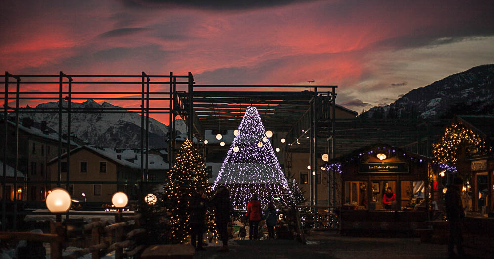 Menschen bestaunen die Christbäume und Beleuchtung auf dem Weihnachtsmarkt in Aosta, Italien, dahinter der Sonnenuntergang