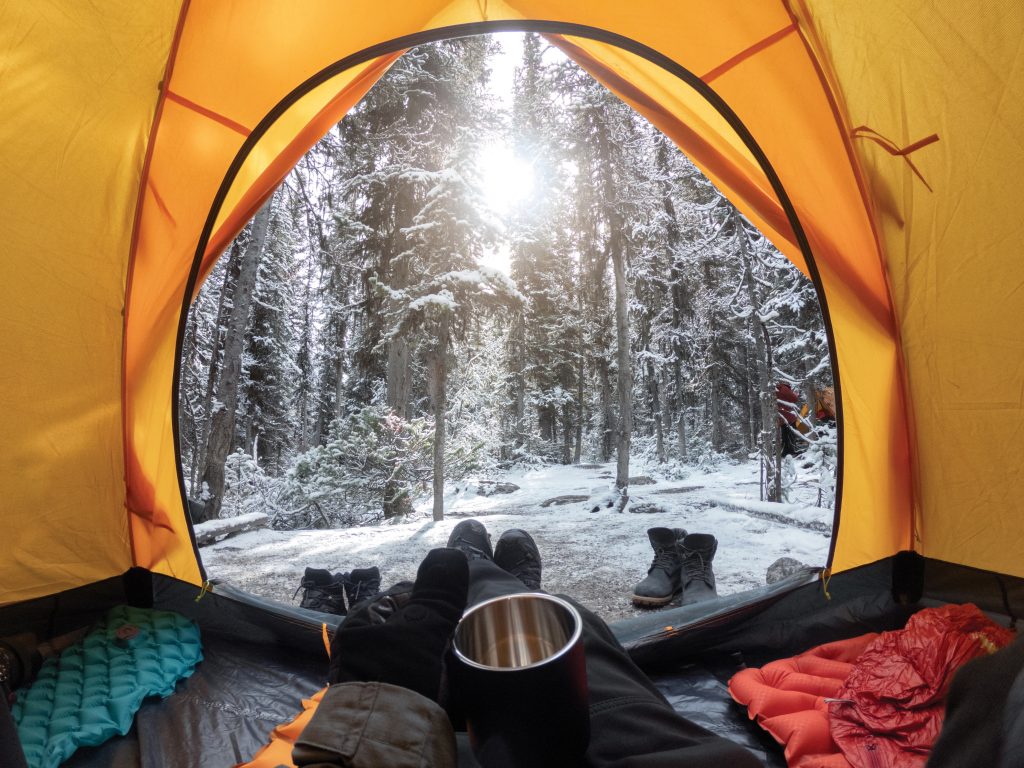 Blick aus einem orangefarbenen Zelt auf eine winterliche Waldlandschaft, behandschuhte Hände halten einen Thermosbecher, Wintercamping, Urlaub im Schnee