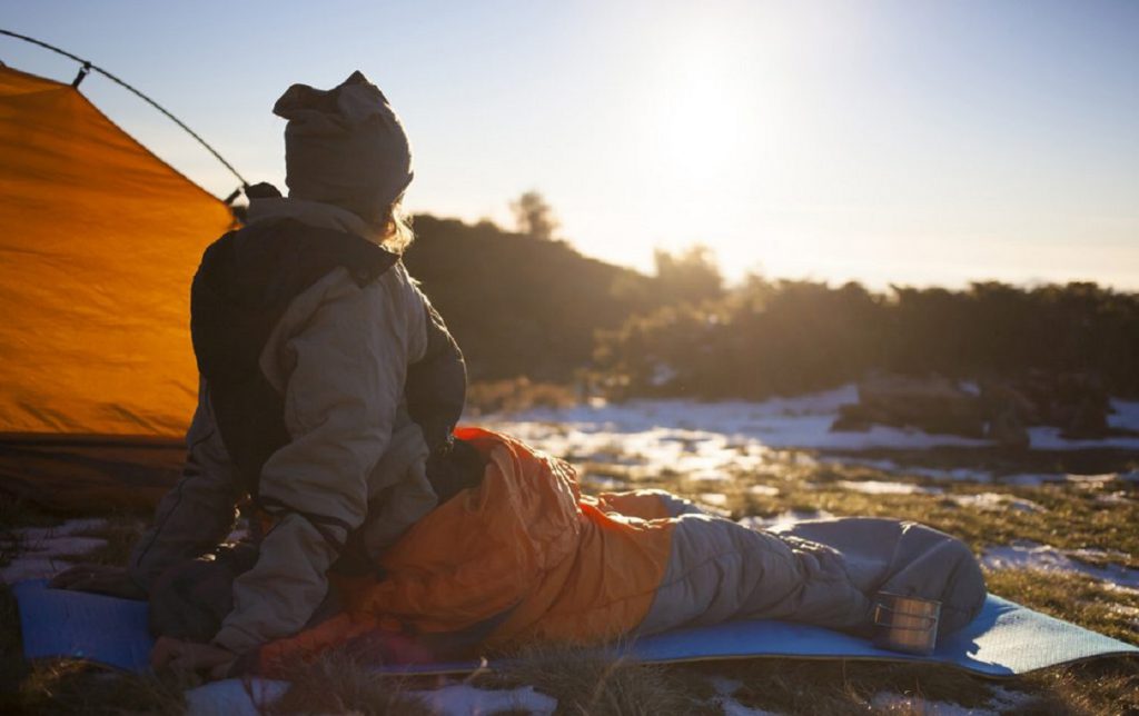 Person im Schlafsack vor ihrem Zelt, winterliche Landschaft mit Schnee und Sonne