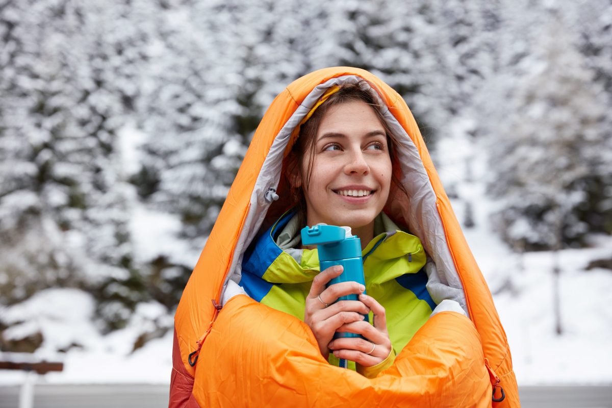 Lächelnde Frau im orangenen Schlafsack vor einer winterlichen Landschaft mit Thermosflasche in der Hand