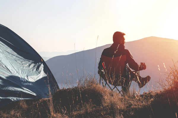 Mann sitzt neben seinem Zelt auf einem Campingstuhl und blickt auf einen Berg