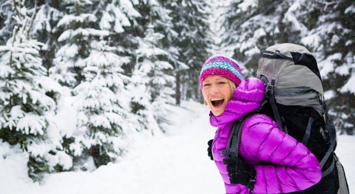 Frau mit lilafarbener Jacke, Wollmütze und Rucksack lacht in die Kamera, im Hintergrund verschneite Waldlandschaft