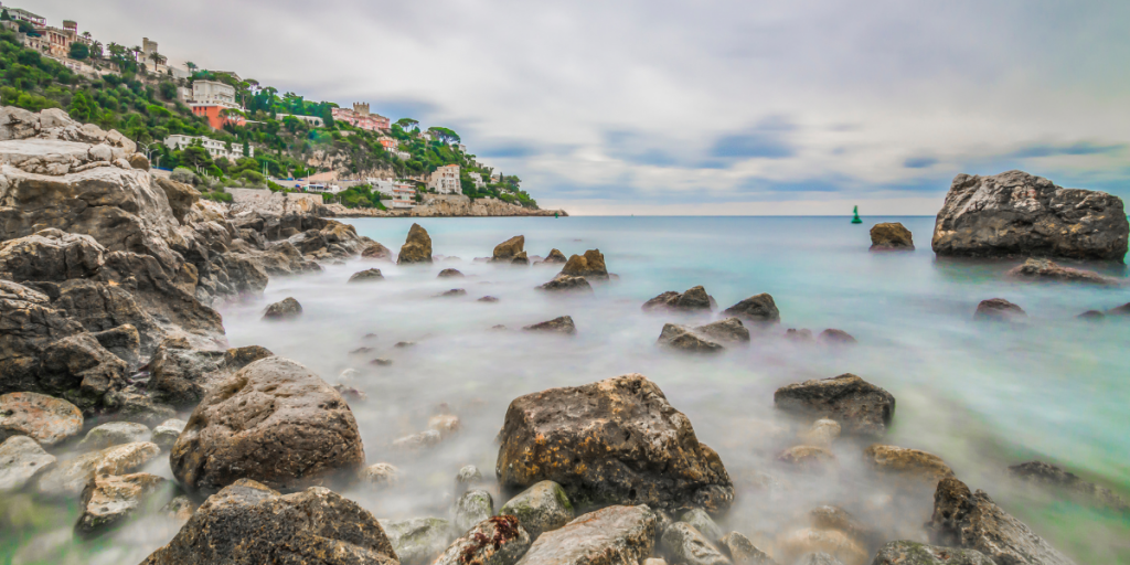 Felsen im Wasser vor der Côte d’Azur in Südfrankreich, im Hintergrund ein Dorf auf den Klippen