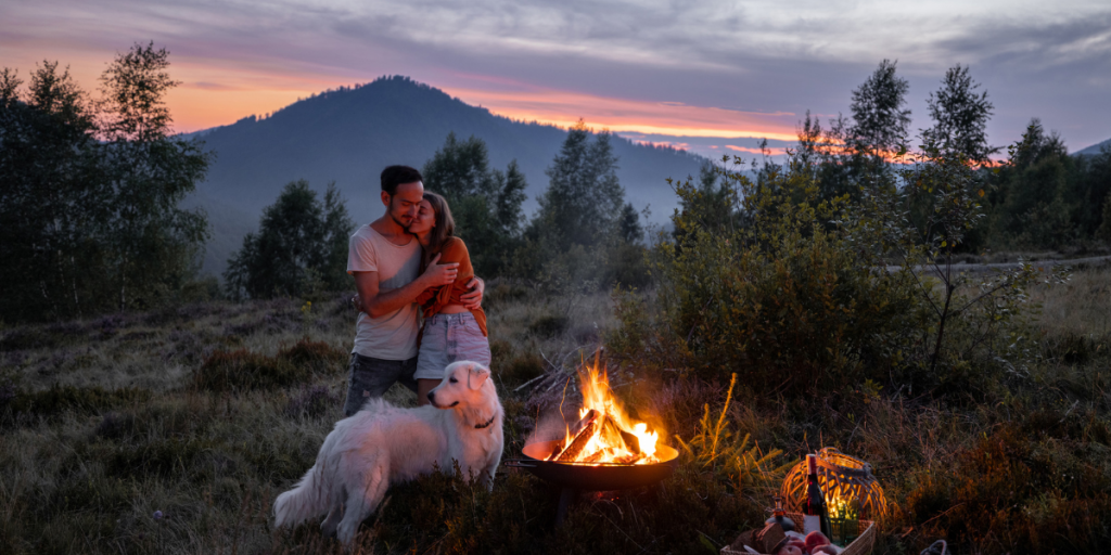 Paar, sich umarmend, mit Hund vor einem Lagerfeuer, im Hintergrund Sonnenuntergang hinter Berg- und Waldlandschaft