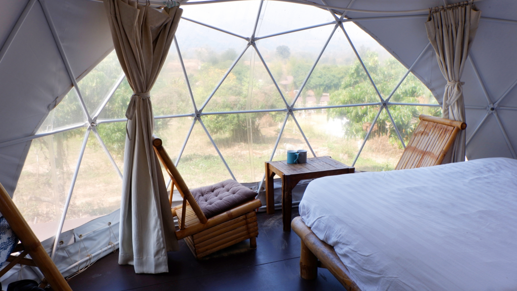 Blick aus einem durchsichtigem Glamping-Pod mit Bett, Sessel und Tisch im Innern, außen Landschaft mit Wald