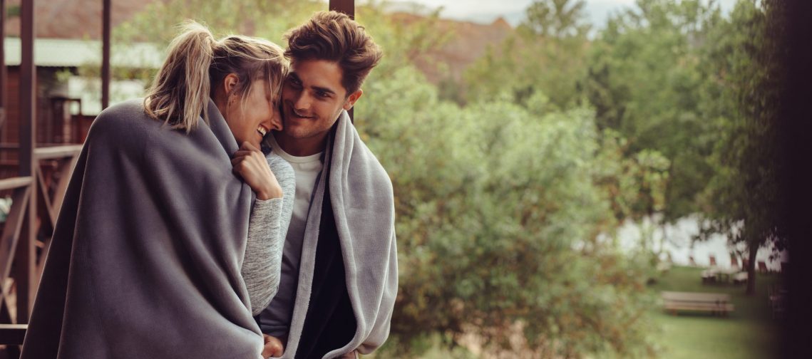 Paar umarmt sich glücklich lächelnd in eine Decke eingewickelt, im Hintergrund Waldlandschaft