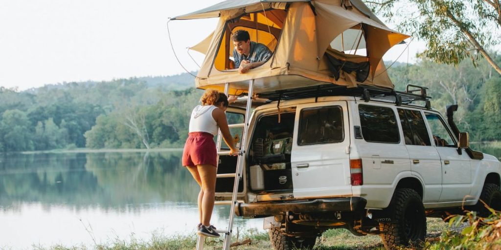 Paar beim Camping am See, Frau steht auf Leiter hinter Campervan, Mann im Zelt